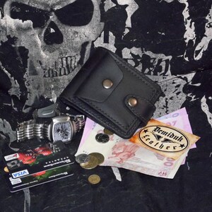 May include: A black leather wallet with two snap closures, a silver wristwatch, credit cards, coins, and paper currency. The wallet is rectangular and has a simple design. The watch has a silver band and a square face. The image also includes a Demiduk Leather tag.