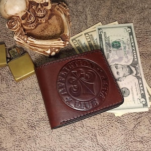 May include: A brown leather wallet with a embossed design that says "Rebel With A Cause Live Free". The wallet is closed and sitting on a table with some US dollar bills.