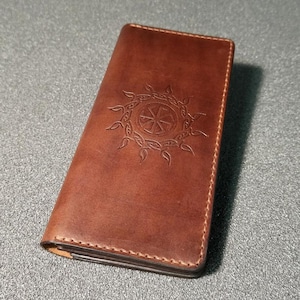May include: A brown leather wallet with an embossed sun symbol. The rectangular wallet has visible stitching around the edges. The leather has a warm, rich tone. The wallet's dimensions are approximately 20 cm in length.