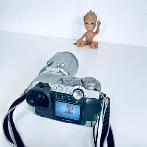 May include: A silver and black Sony camcorder with a black and white striped strap. The screen displays a baby image. A small brown Groot figurine is in the background, waving.