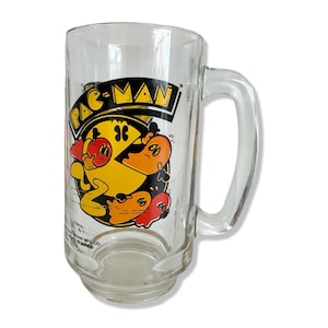 May include: A clear glass Pac-Man mug with a yellow Pac-Man character and ghosts in red, orange, blue, and pink. The mug has a handle and says "Pac-Man" in black letters.