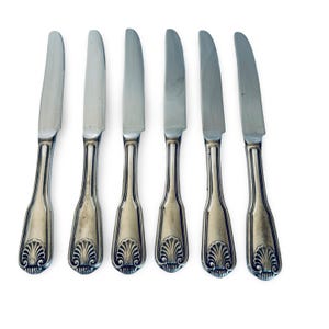 May include: A set of six silver-toned metal dinner knives arranged in a vertical row. Each knife features a smooth, polished blade and a decorative handle with a prominent shell-shaped motif at the base and vertical grooved detailing along the stem.
