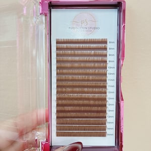 May include: A pink plastic case containing rows of brown eyelash extensions arranged by length. The white backing features the text Perfection Studio Lash & Brow and labels for lengths ranging from 8mm to 14mm printed next to each row of lashes.