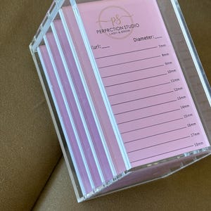Könnte beinhalten: Ein transparenter Acrylorganizer mit rosa Karten für Wimpernextensions. Die Karten enthalten eine Liste von Wimperngrößen in Millimetern, von 7 mm bis 18 mm. Der Organizer ist mit "Perfection Studio Lash & Brow" beschriftet.