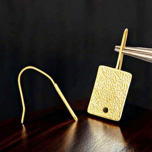 May include: A pair of gold-colored earrings. Each earring features a textured rectangular pendant with a small hole and a curved hook. The earrings are displayed on a dark wooden surface.
