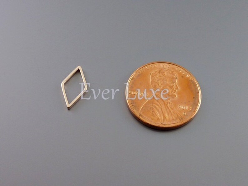 May include: A small, gold-colored, open diamond-shaped charm next to a penny. The charm is a simple geometric design, and the penny provides a size reference. The background is a solid gray.