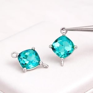 May include: Two square-cut, teal-colored gemstone charms with silver-colored metal settings. Each charm has a loop at the top and a small loop at the bottom. One charm is held by a pair of tweezers.