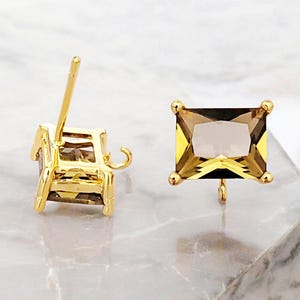May include: Two gold-colored stud earrings with rectangular, light brown gemstones. The earrings have a post and a small loop for attachment. The gemstones are set in a gold-colored square frame with small gold-colored balls at each corner.