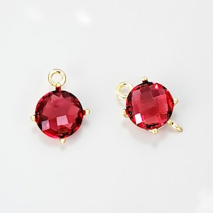 May include: Two red, faceted, round gemstone charms with gold-colored metal findings. Each charm has a loop at the top for hanging and small decorative prongs around the gemstone. The gemstones are a deep, rich red color.