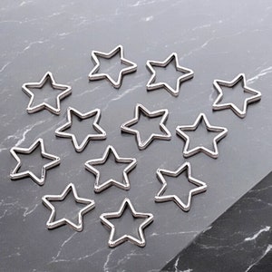 Può includere: Un set di 12 ciondoli in metallo a forma di stella argentati. I ciondoli sono tutti della stessa dimensione e forma, e hanno una finitura liscia e lucida.