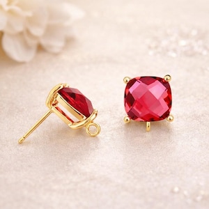 May include: A pair of gold-colored stud earrings with large, square, faceted, ruby-red gemstones. The earrings have a post and a small loop for attaching a backing. The gemstones are set in a gold-colored metal setting.
