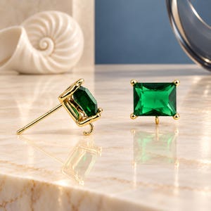 May include: A pair of gold stud earrings featuring rectangular emerald green gemstones. Each earring has a four-prong setting and a small loop at the bottom for attaching additional charms or dangles. The posts are thin and metallic gold.