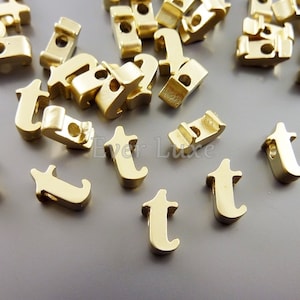 May include: A group of gold-colored metal letter beads in the shape of the letter "T". The beads have a hole in the center for stringing.