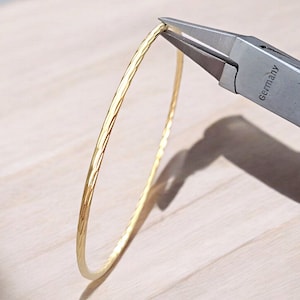 May include: A gold bangle bracelet with a textured surface is being opened with pliers. The pliers are labeled "Germany".
