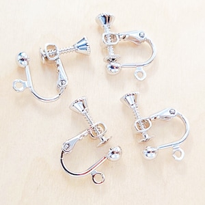 May include: Close-up of five silver-colored clip-on earring findings. Each finding has a screw-back mechanism, a curved clip, and a small loop. The findings are arranged on a light-colored wooden surface.