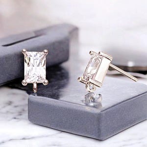 Op de afbeelding: Twee zilverkleurige oorstekers met rechthoekige, heldere, facetgeslepen stenen. Elke oorbel heeft een pin voor piercing en een kleine lus aan de onderkant. De oorbellen worden gepresenteerd op een grijze ondergrond.