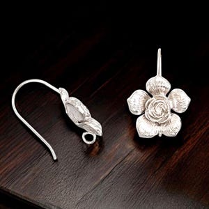 May include: A pair of silver-colored flower earrings. One earring is a complete flower with five petals and a central rose design. The other earring has a hook and a leaf-shaped design. The earrings are on a dark wooden surface.