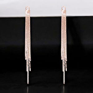 Peut inclure: Une paire de boucles d'oreilles à franges en or rose. Chaque boucle d'oreille est composée d'une boucle supérieure et de plusieurs brins de chaîne délicate qui descendent en cascade. Les boucles d'oreilles ont un aspect texturé et sont suspendues sur un fond noir.