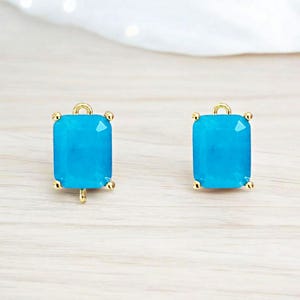 May include: Two rectangular turquoise-colored gemstone pendants with gold-colored accents. Each pendant has a gold loop at the top and four gold prongs at the corners. The pendants are displayed on a light-colored wooden surface.