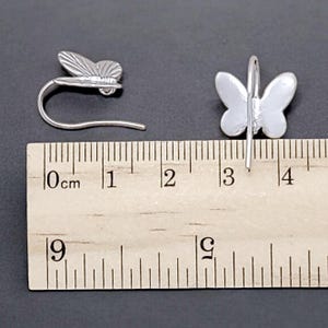 May include: Two silver butterfly earrings with a hook closure. The earrings are measured against a ruler, showing they are approximately 1.5 inches long.