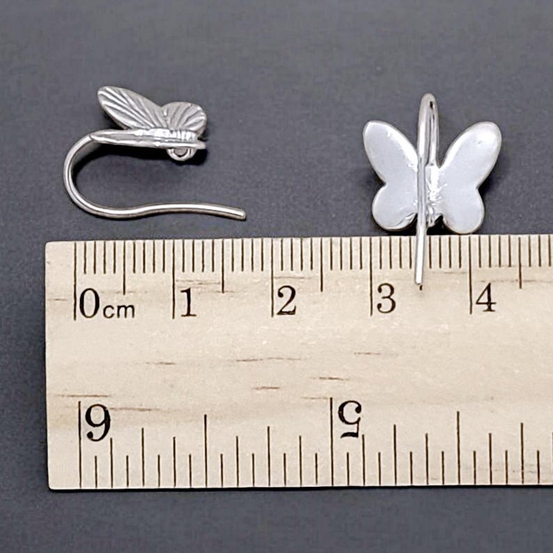 May include: Two silver butterfly earrings with a hook closure. The earrings are measured against a ruler, showing they are approximately 1.5 inches long.