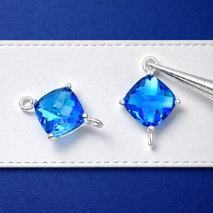 May include: Two square, faceted, bright blue gemstone pendants with silver-colored metal findings. The gemstones are set with small silver prongs at each corner. One pendant is being held by tweezers. The background is white and blue.