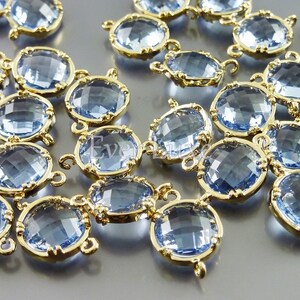Puede incluir: Un grupo de conectores de cristal azul claro con engastes de metal dorado. Los conectores tienen forma redonda y están diseñados para la elaboración de joyas.