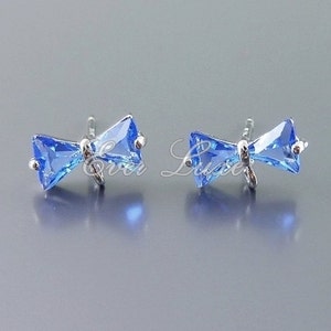 May include: A pair of bow-shaped stud earrings. Each earring features a light blue, rectangular-cut gemstone set in a silver-toned metal. The bow design is detailed and elegant, with a post for secure wear.