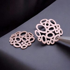 Peut inclure: Une paire de boucles d'oreilles en or rose avec un design délicat et complexe de cercles qui se chevauchent. Les boucles d'oreilles sont en métal et ont une finition polie.
