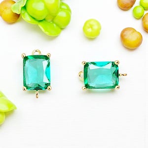 May include: Two rectangular, emerald-green gemstone charms with gold-colored settings. Each charm has a loop at the top and a small loop at the bottom for attaching to jewelry. The gemstones are faceted and have a clear, transparent appearance.