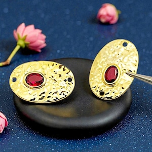 May include: Two gold-colored oval earrings with a hammered texture, each featuring a central, oval-shaped, deep red gemstone. The earrings have small holes at each end. The earrings are displayed on a black surface with pink flowers in the background.
