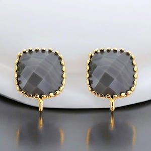 May include: A pair of gold-tone clip-on earrings with a square-shaped gray gemstone. The earrings have a beaded edge and a simple clip closure.