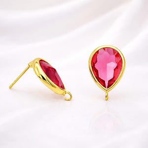 May include: Two gold-colored stud earrings with a teardrop-shaped, faceted, red gemstone. The gemstones are set in a gold bezel. The earrings have a post and a small loop at the bottom.