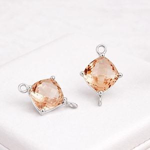 May include: Two square-cut, peach-colored gemstone charms with silver-tone metal settings. Each charm has a loop at the top and a small loop or post at the bottom for attaching to jewelry. The gems are faceted, reflecting light.