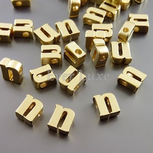 May include: A group of gold letter beads, all in the shape of the letter "n". The beads are scattered on a gray surface.
