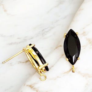 May include: A pair of gold-colored earrings with a black, marquise-shaped stone. The earrings have a post and a small loop at the bottom. The background is a white and gray marble pattern.