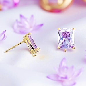 May include: A pair of gold-colored stud earrings with rectangular, light purple gemstones. The earrings are set against a soft, white background with blurred purple flower petals.