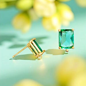 May include: A pair of gold-colored stud earrings with emerald-cut, green gemstones. The earrings are set in a gold-colored metal and have a post and butterfly backing. The background is a soft, light blue with blurred yellow and green accents.