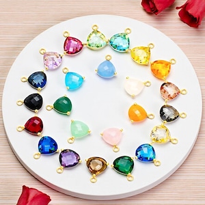 May include: A collection of colorful gemstone pendants in various shapes, including triangles and teardrops, with gold-colored loops for attaching to jewelry. The gemstones are arranged on a white circular surface.