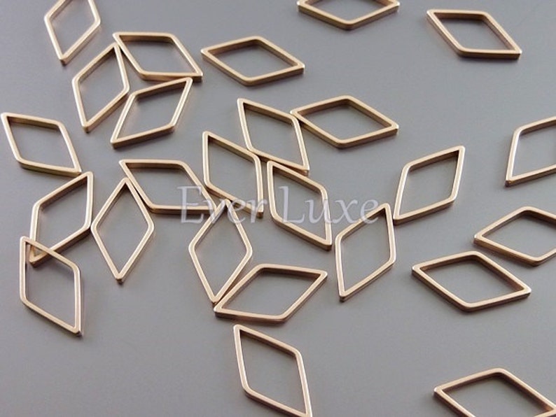 May include: A collection of small, gold-colored diamond-shaped metal frames scattered on a gray surface. The frames are open in the center and have a smooth, polished finish. These components could be used for jewelry making or other craft projects.