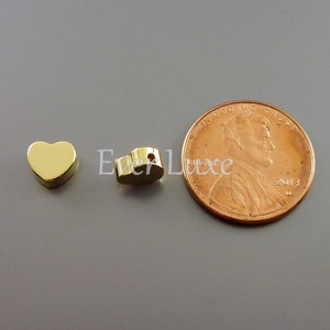 May include: Two gold-colored heart-shaped charms, one with a hole, next to a US penny for size comparison.