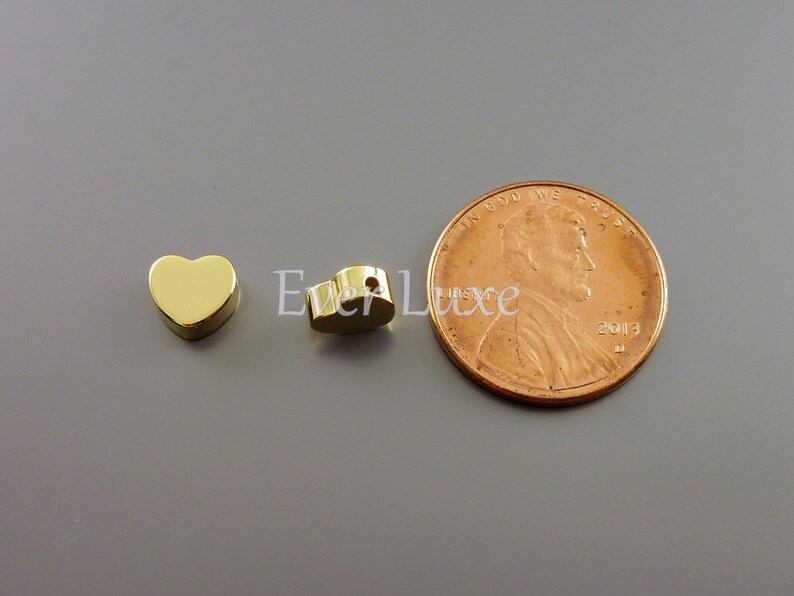 May include: Two gold-colored heart-shaped charms, one with a hole, next to a US penny for size comparison.