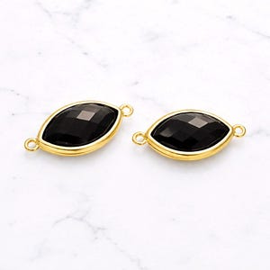 May include: Two black marquise-shaped gemstone beads with gold-colored bezels and small loops on each end. The faceted black stones are set in a gold-tone metal. The beads are displayed on a white marble surface.