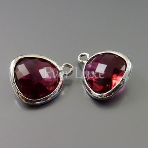 Pode incluir: Dois pingentes prateados com um cristal facetado em forma de lágrima de cor vermelho escuro.