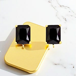 May include: A pair of black rectangular gemstone earrings with gold-colored settings. The earrings are displayed on a gold-colored rectangular surface against a white background. The gemstones have a faceted appearance.