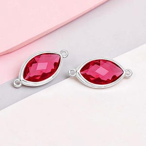 May include: Two marquise-shaped jewelry components with a vibrant red gemstone center and a silver-colored metal frame. Each component has a loop on either end for attachment, ideal for jewelry making. The gemstones have a faceted cut, reflecting light.