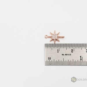 Op de afbeelding: Een ros&eacute;gouden sterbedel met heldere zirkonia-stenen. De bedel is ongeveer 2,5 cm lang en wordt naast een liniaal getoond ter referentie van de grootte.