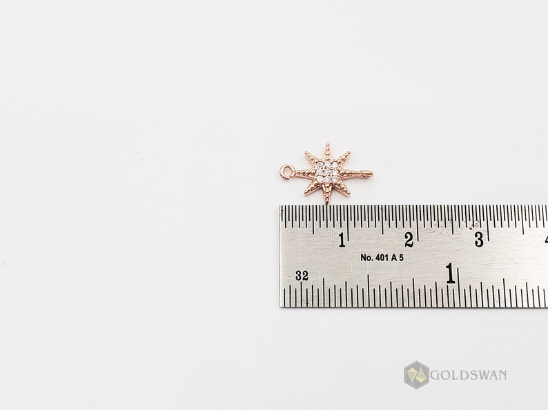 Op de afbeelding: Een ros&eacute;gouden sterbedel met heldere zirkonia-stenen. De bedel is ongeveer 2,5 cm lang en wordt naast een liniaal getoond ter referentie van de grootte.