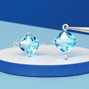 May include: Two square-cut, light blue gemstone earrings with silver-colored metal settings. Each earring has a loop for attachment and small decorative accents. The earrings are displayed on a white and blue surface.