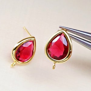 May include: A pair of gold-tone earrings with red teardrop-shaped crystals. The earrings have a simple design and are perfect for everyday wear.